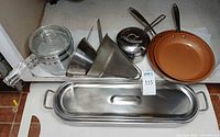 All items laid out: Pyrex double boiler, fish poacher with lid, mesh strainer, conical strainer, stainless saucepan, two copper-tone fry pans