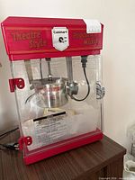 Front view of red Cuisinart CPM-25C popcorn maker showing clear doors, stainless steel kettle, power cord and manual inside