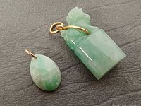 Carved cylindrical jadeite pendant and faceted oval jadeite pendant with gold hoops on grey background