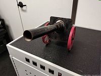 Side view of metal toy cannon showing black barrel, red wheels, surface rust