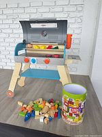 Full view of Melissa & Doug toy grill with play food pieces and tub of wooden blocks