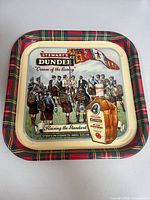 Front of square tartan-border tray featuring Highland parade illustration and whisky bottle