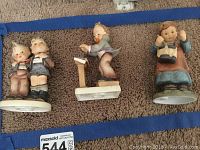 Three Goebel porcelain figurines displayed upright on carpet. The figurines include two boys standing together, a boy playing a drum, and a girl holding a basket.