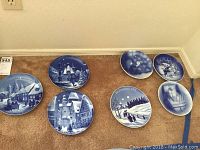 Ten collectible blue and white Christmas-themed porcelain plates arranged on carpet. Includes multiple designs featuring winter village scenes, children, and festive imagery.