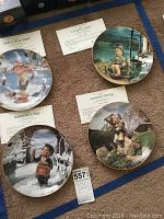 Four M.I. Hummel collector plates displayed flat on carpet, titles visible along with COA cards: Valentine's Day, Autumn Outing, Carefree Days, Year's First Snow.