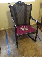 Full view of vintage wooden rocking chair showing cane back, carved dark wood frame, and floral embroidered red seat.