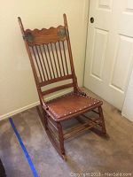 Full front and side angle showing the wooden rocking chair with spindle back, seat with metal studs, and rocker legs.
