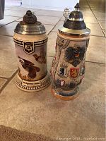 Two German beer steins on a tiled floor, one with bird artwork, the other decorated with heraldic shields