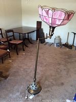 Full view of the brass floor lamp with pink Tiffany style shade, showing the lamp standing upright on carpeted floor surrounded by chairs and other household items.
