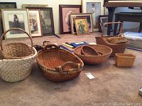 Group of 7 baskets on carpet floor with framed pictures in background. Various sizes and shapes, including oval and rectangular with handles.