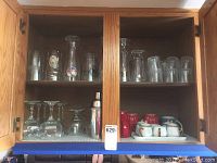 Glass cabinet with multiple shelves, displaying different glassware including beer glasses with logos, tall clear glasses, wine glasses, brandy snifters, a metal cocktail shaker, and ceramic mugs in red and blue with saucers.