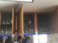 Three wooden kitchen cabinets with glassware inside. Left and right cabinet doors open showing various glassware including short glasses, stemmed glasses, tall glasses, and metal cordial glasses on shelves.