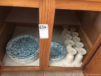 View of stacked blue mottled plates, blue mottled bowls, and white vitrified China mugs stored inside a wooden cabinet.