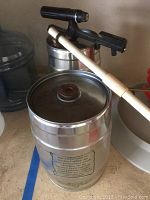 Photo showing two metal mini kegs with printed instructions, plastic beer tap attached to one, and a plastic spoon resting on keg lid.