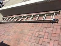 Full view of a long, metal extension ladder laid horizontally on brick pavement, showing general wear and dirt.