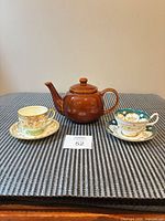 Overall lot view: Brown Betty teapot centered between two vintage teacup-saucer sets