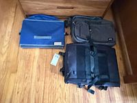 Three items laid out: black backpack, grey Solo briefcase, blue Costco divider bin