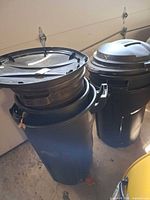 Two of the black plastic trash cans with lids stacked, showing handles and scuffs