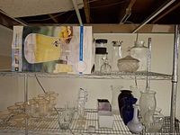Overall view of two wire shelves showing boxed beverage dispenser, pitchers, ribbed jar, candy dish, vases, tumblers and glasses