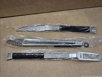 Three stainless steel BBQ tools—knife, tongs, spatula—wrapped in plastic