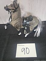 Full left side view showing dapple grey body, white mane and tail, black saddle, bridle, reins, metal-tone stirrups