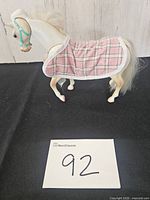 Left side view of white plastic toy horse wearing pink plaid blanket