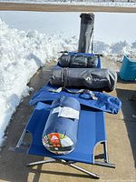 Lineup of camping gear: cot, sleeping bag, rolled pads, gear bags, chair