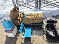 Overall view of lot showing cot, bedrolls, cooler, water jug, chair, grill and bag