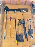 All tools hanging on wall including landscape rake, leaf rake, shovel, pickaxe, telescopic pole