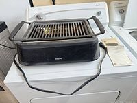 Front view of Philips grill on top of washing machine showing housing, grate, handles, power cord