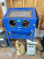 Front view of blue sandblasting cabinet on stand with two glove ports, upper window, lower shelf, media bags below
