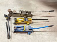 Three trolley jacks with handles laid on floor (silver, yellow, blue)