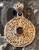 Front view of round sterling silver pendant showing scroll filigree, cross bars and central red stone