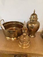 Three brass pieces grouped: handled planter bucket, lidded ginger jar urn, candlestick form