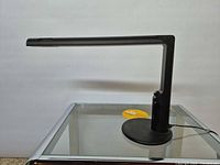 Side profile of black Abele desk lamp on glass table showing right-angle arm and base