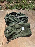 Two olive drab US military duffel bags on concrete, one large side-zip duffel, one top-load sea bag