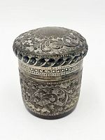 Side view of ornate cylindrical silver spice box with lid showing repousse work