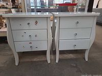 Front view of both white gloss nightstands showing three drawers, curved legs and sticker residue on one unit