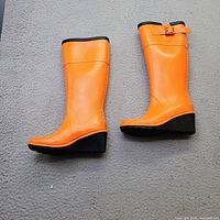 Both orange Michael Kors rain boots laid flat showing outer sides, wedge heels and buckle detail
