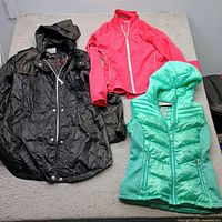 Three outerwear pieces laid out: black hooded jacket, pink jacket, teal vest