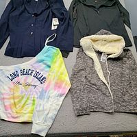 Four new women’s size L garments laid out: navy blazer, dark grey quarter-zip, pastel tie-dye sweatshirt, grey sherpa hoodie with tags visible