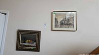 Both framed artworks hung on wall: still life lower left, cityscape upper right