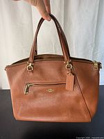 Front view of Coach satchel showing logo, tag, front zip pocket, handles, gold hardware