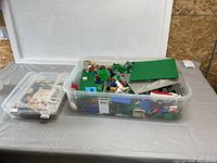 Large clear tote filled with mixed LEGO pieces next to smaller bin of manuals