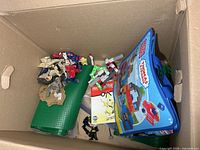 Box containing green mats, loose Mega Bloks pieces, DS case, micro figures, Thomas bag