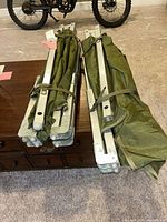 Two folded olive green camp cots with aluminum frames strapped together