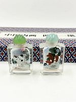 Front view of two snuff bottles showing painted scenes and stoppers, case blurred behind