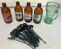 Group shot of all items: four amber beer bottles, green Coca-Cola mug, pile of black stir sticks