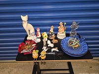 Overall lot on black table, showing tall cat figurine, red glass bowl, assorted porcelain figurines, blue bowl with glass rings