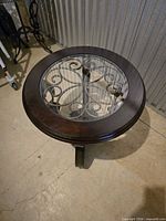 Full view of round side table showing wood rim, glass top with scroll design, and iron base